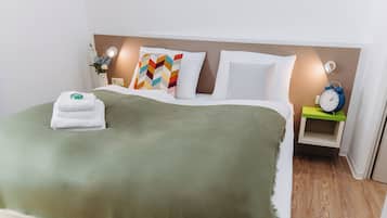 A neatly made bed with a green blanket, white pillows, and a colorful pillow. There is a small bedside table with a clock and a vase with flowers.