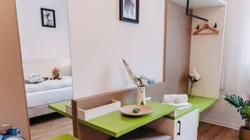 Habitación de hotel con un escritorio verde, una cama con un osito de peluche, un espejo, un reloj en la pared y un jarrón con flores.