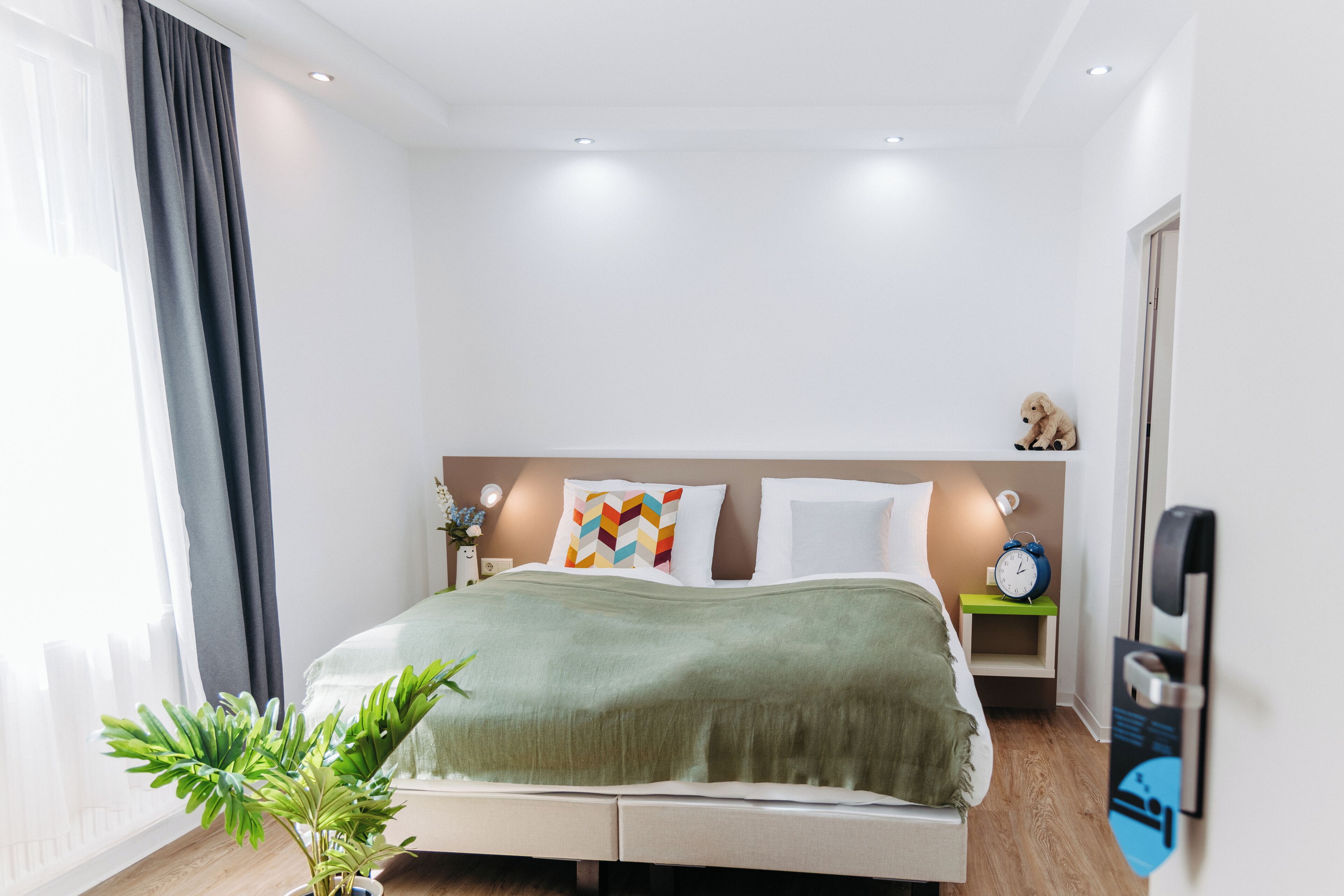 Una habitación de hotel moderna con una cama bien hecha, una mesita de noche con lámpara, un reloj y una planta pequeña.