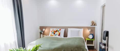 Una habitación de hotel moderna con una cama bien hecha, una mesita de noche con lámpara, un reloj y una planta pequeña.