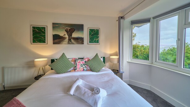 A hotel room with a large bed, a view of greenery through the window, and artwork on the wall.
