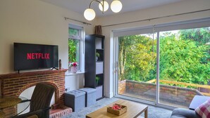A living room with a fireplace, a television displaying 'NETFLIX', a sofa, a coffee table, and a view of greenery through large windows.