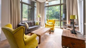 Una sala de estar moderna con un sofá, un sillón amarillo y una mesa de centro de madera. Amplios ventanales ofrecen vistas al verdor exterior.