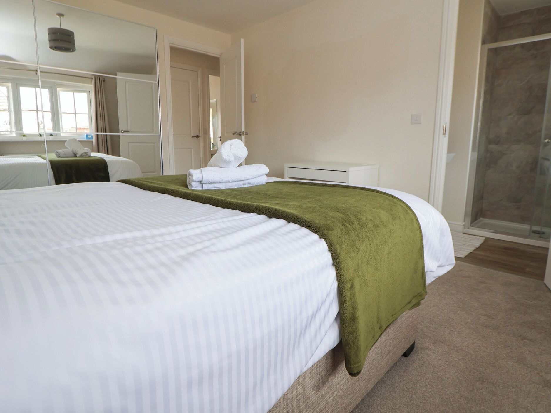 A hotel room with a large bed, a green throw blanket, a white towel, a small bedside table, a mirror, and a shower area.