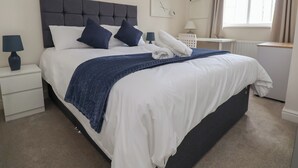 Una cama bien hecha con cabecera oscura, ropa de cama blanca y dos almohadas oscuras. Una manta azul está doblada al pie de la cama. Se ve una mesita de noche blanca con una lámpara y una pequeña silla blanca. Una ventana con cortinas se encuentra a la derecha de la cama.