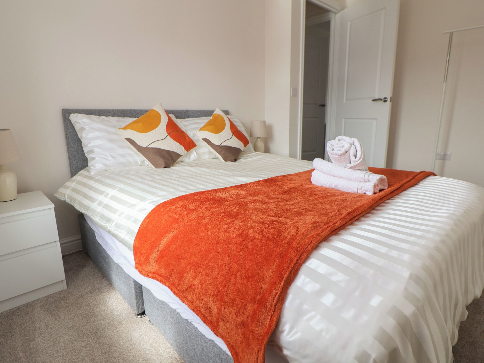 A neatly made bed with white and orange bedding, a grey headboard, and a red throw blanket.