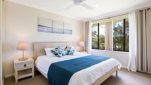 A bedroom with a large bed, two bedside tables, a ceiling fan, a window with curtains, and a painting on the wall.