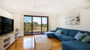 A modern living room with a large flat-screen TV, a blue sofa, a wooden coffee table, and a balcony with a view of the outdoors.