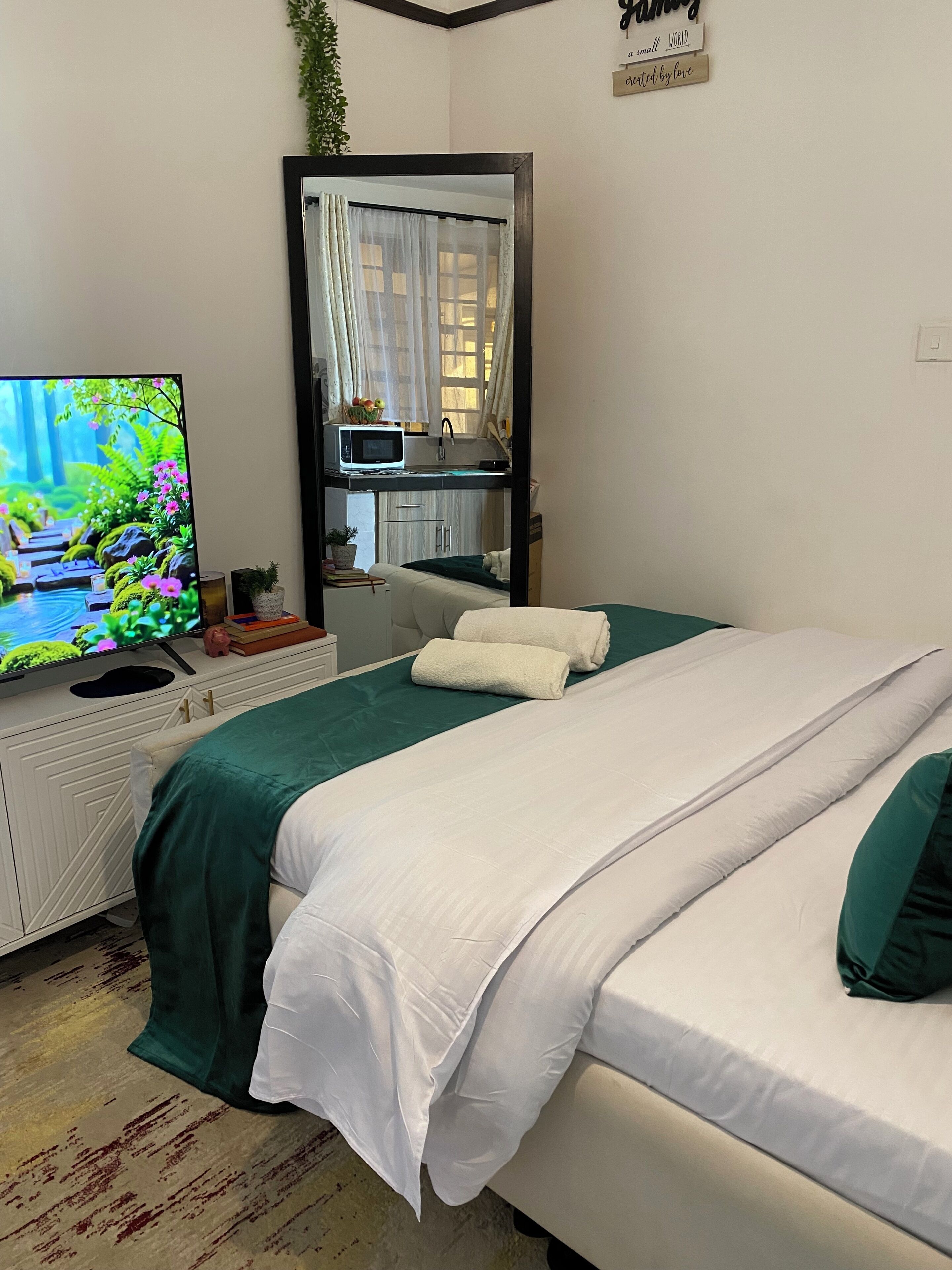 Quarto de hotel com cama, televisão, uma pequena cozinha e janela com cortinas.