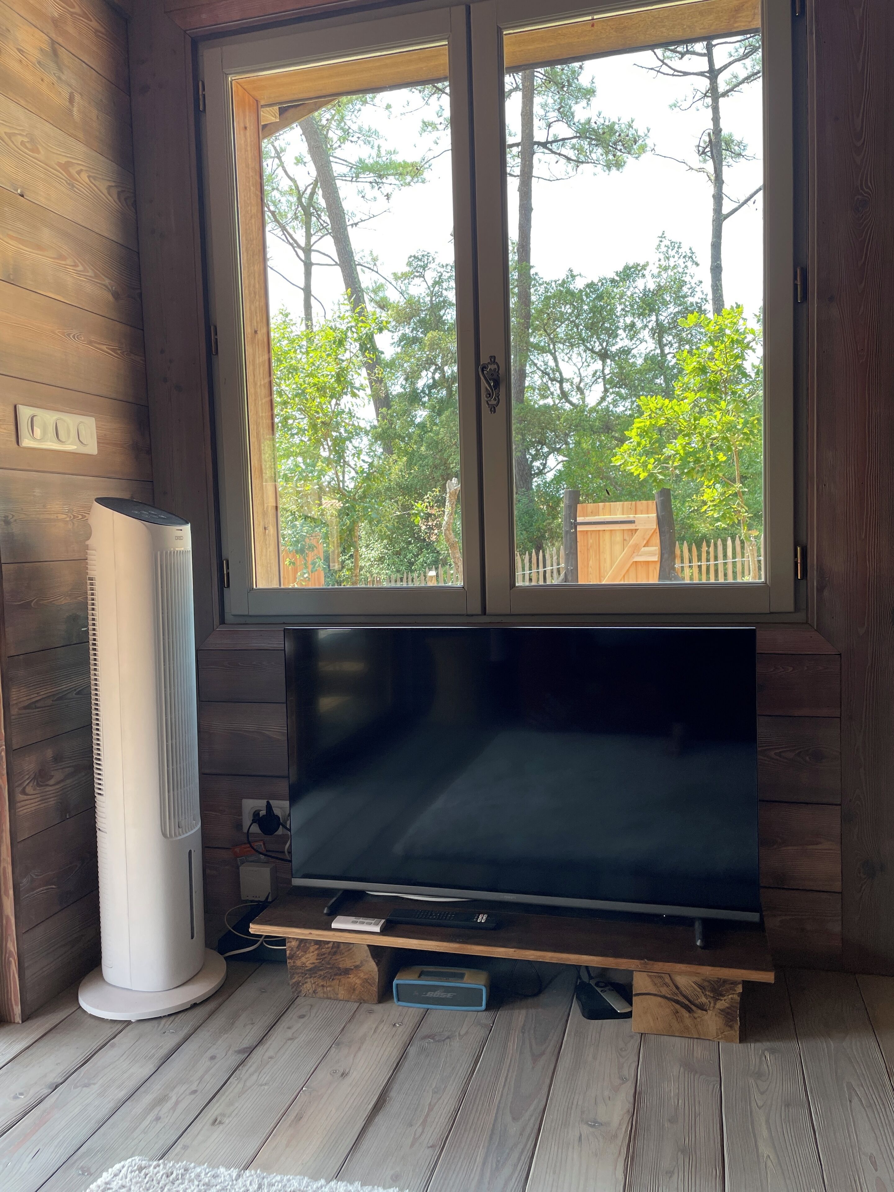 A room with a large flat-screen TV on a wooden stand, a white air conditioner, and a window with a view of trees and a wooden fence.