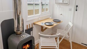 A compact kitchen with a wood-burning stove, a dining table with chairs, and a window with a view of a wooden deck. - Kings Cwtch Shepherd's Hut (Cardiff)