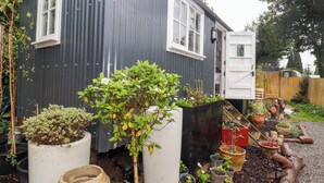 A house with a grey exterior, white windows, and a door. There are various potted plants and a garden area with a gravel path. - Kings Cwtch Shepherd's Hut (Cardiff)