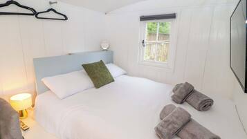 A neatly made bed with white linens and a green pillow, a bedside lamp, a folded towel, and a window with a view of greenery.