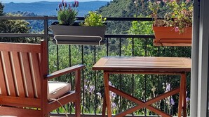 Ein Balkon mit Holzstuhl und Tisch, umgeben von Blumentöpfen und einer malerischen Aussicht auf Hügel und Berge.
