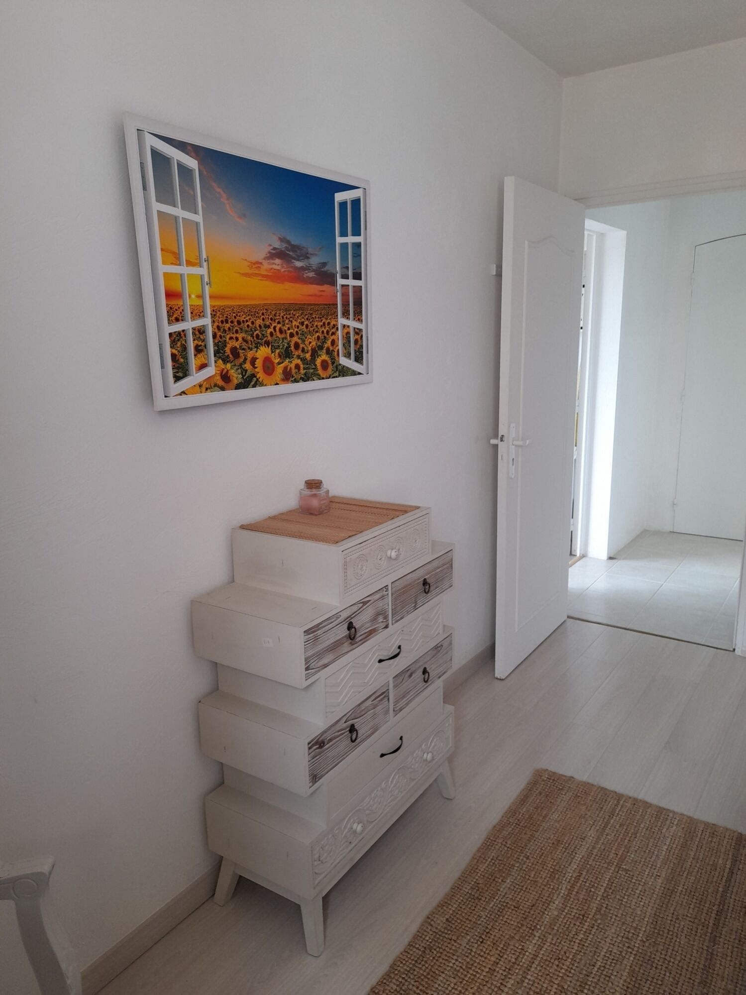 A room with a white wall, a painting of a sunset over a field of sunflowers, a white dresser with drawers, and a door leading to another room.
