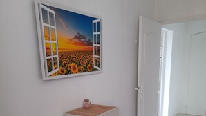 Ein Zimmer mit weißer Wand, einem Gemälde, das einen Sonnenuntergang über einem Sonnenblumenfeld zeigt, einer weißen Kommode mit Schubladen und einer Tür zu einem anderen Raum.