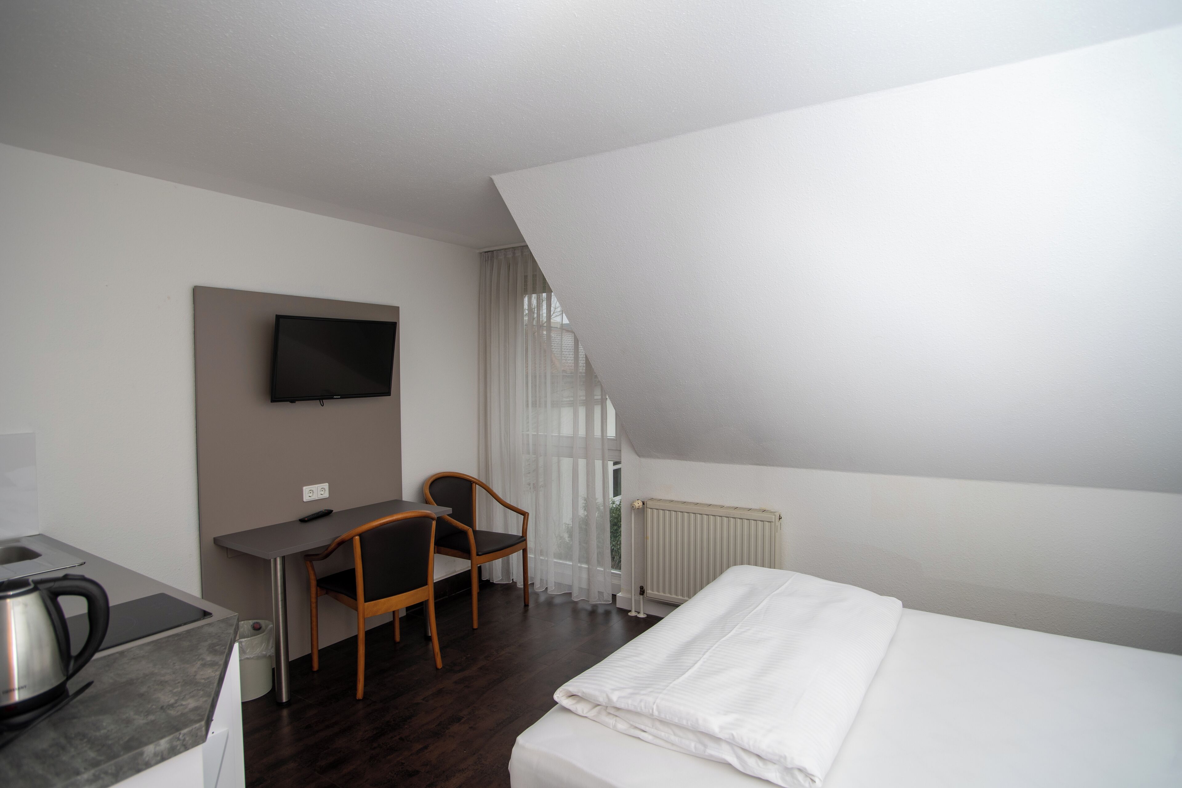 A hotel room with a bed, a desk, a chair, a TV mounted on the wall, and a radiator.