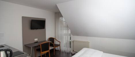 A hotel room with a bed, a desk, a chair, a TV mounted on the wall, and a radiator.