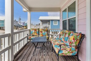 Une véranda couverte avec une terrasse en bois, une table en métal et un canapé coloré agrémenté de coussins à motifs floraux.