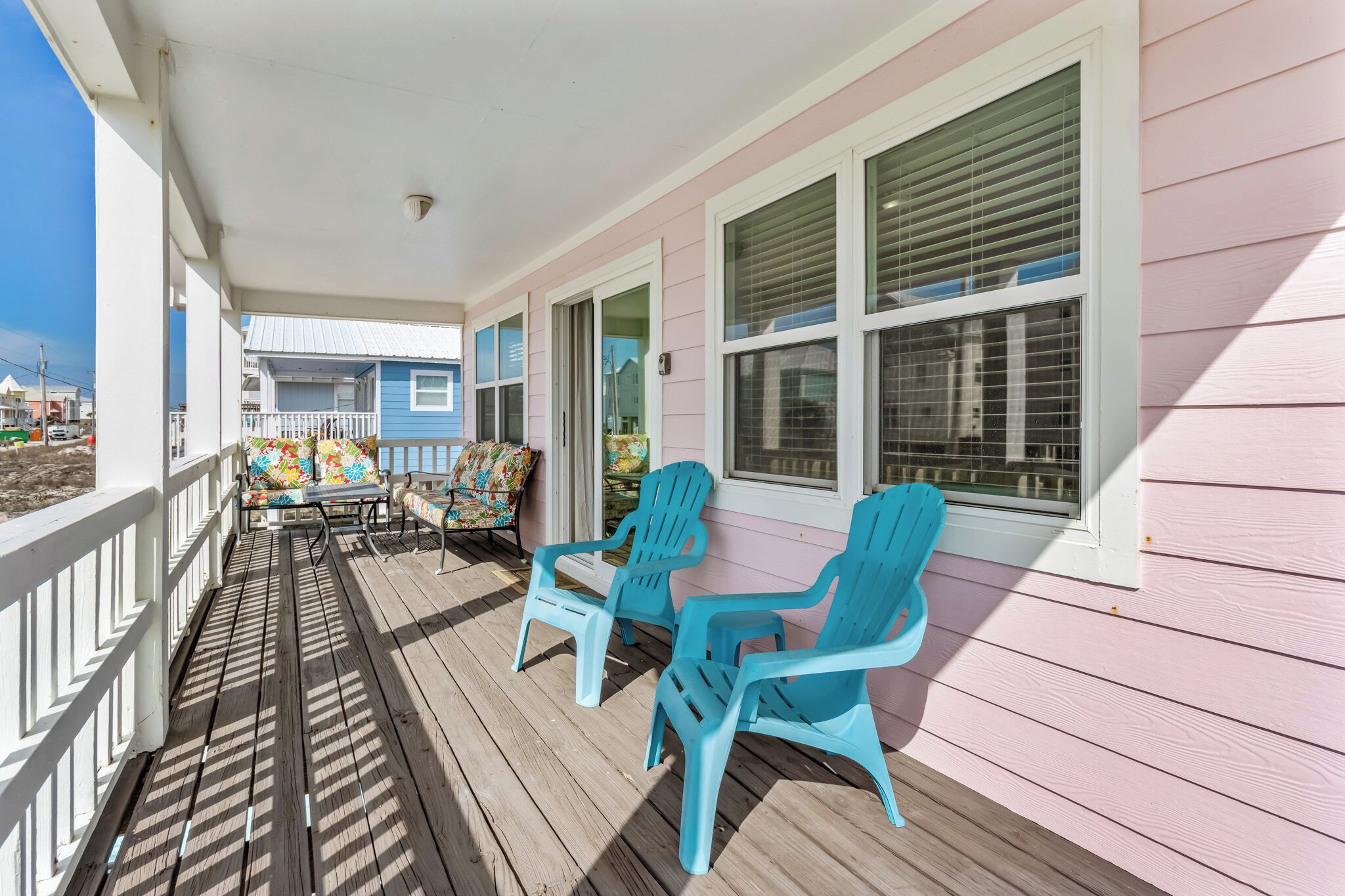 Une véranda couverte avec une terrasse en bois, deux fauteuils Adirondack turquoise, une table avec des coussins colorés et, au loin, une vue sur une maison bleue.