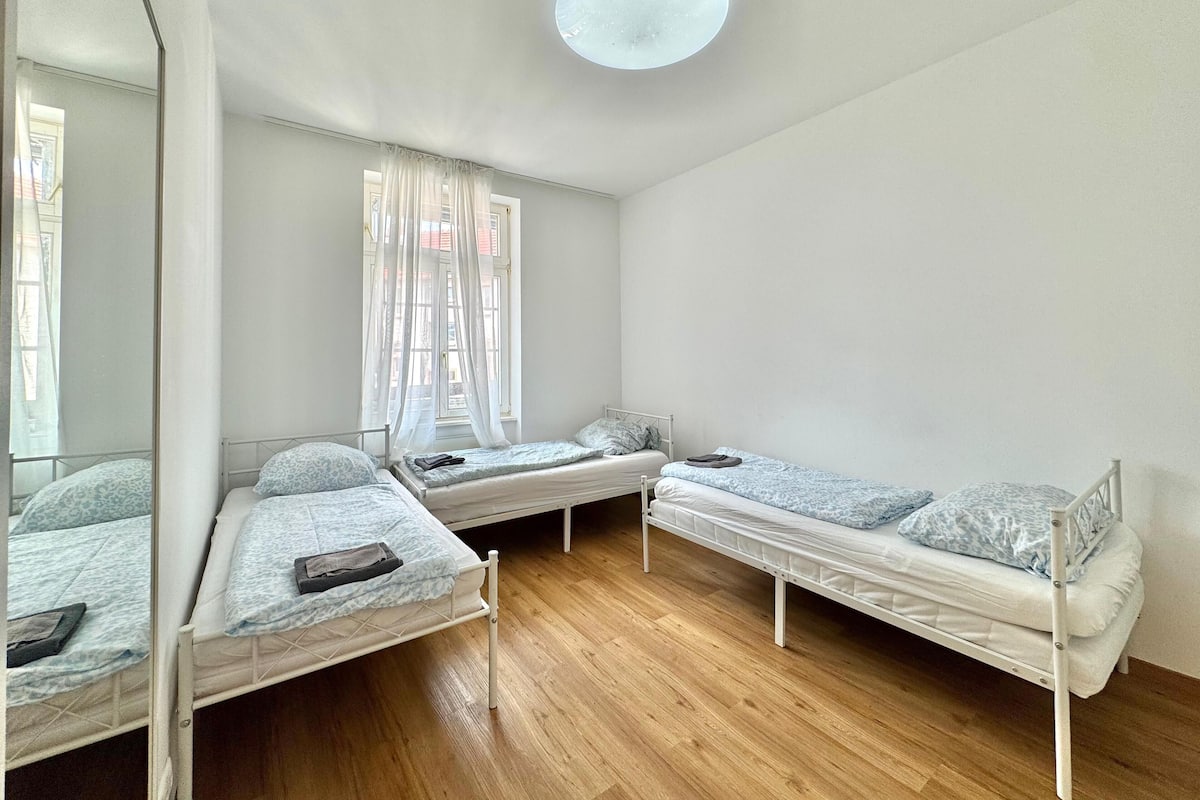 A dormitory room with three single beds, wooden flooring, a large mirror, and a ceiling light.