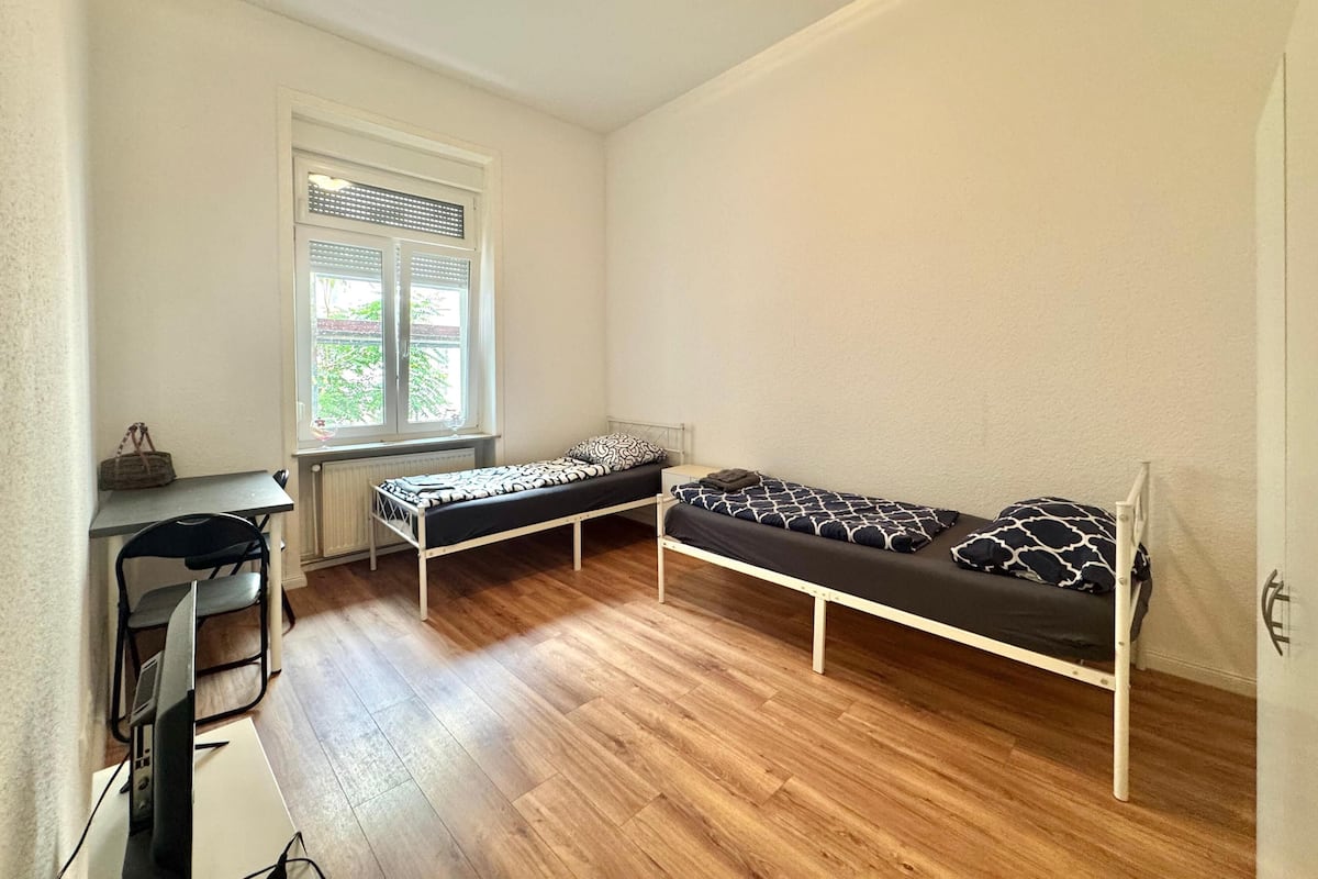 A small, bright room with a wooden floor, two single beds with patterned bedding, a small table, and a chair.