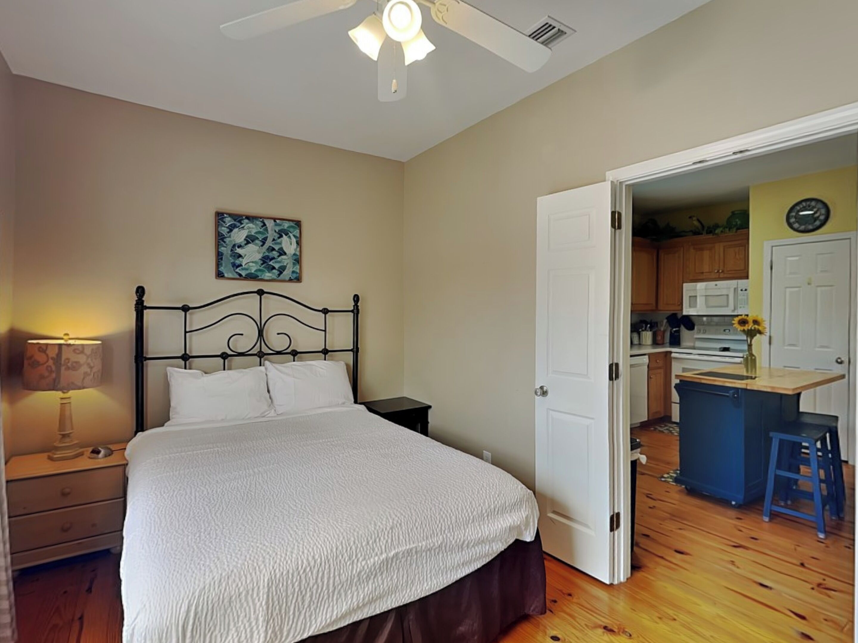 A bedroom with a bed, a nightstand, a lamp, a ceiling fan, and a door leading to a kitchen.