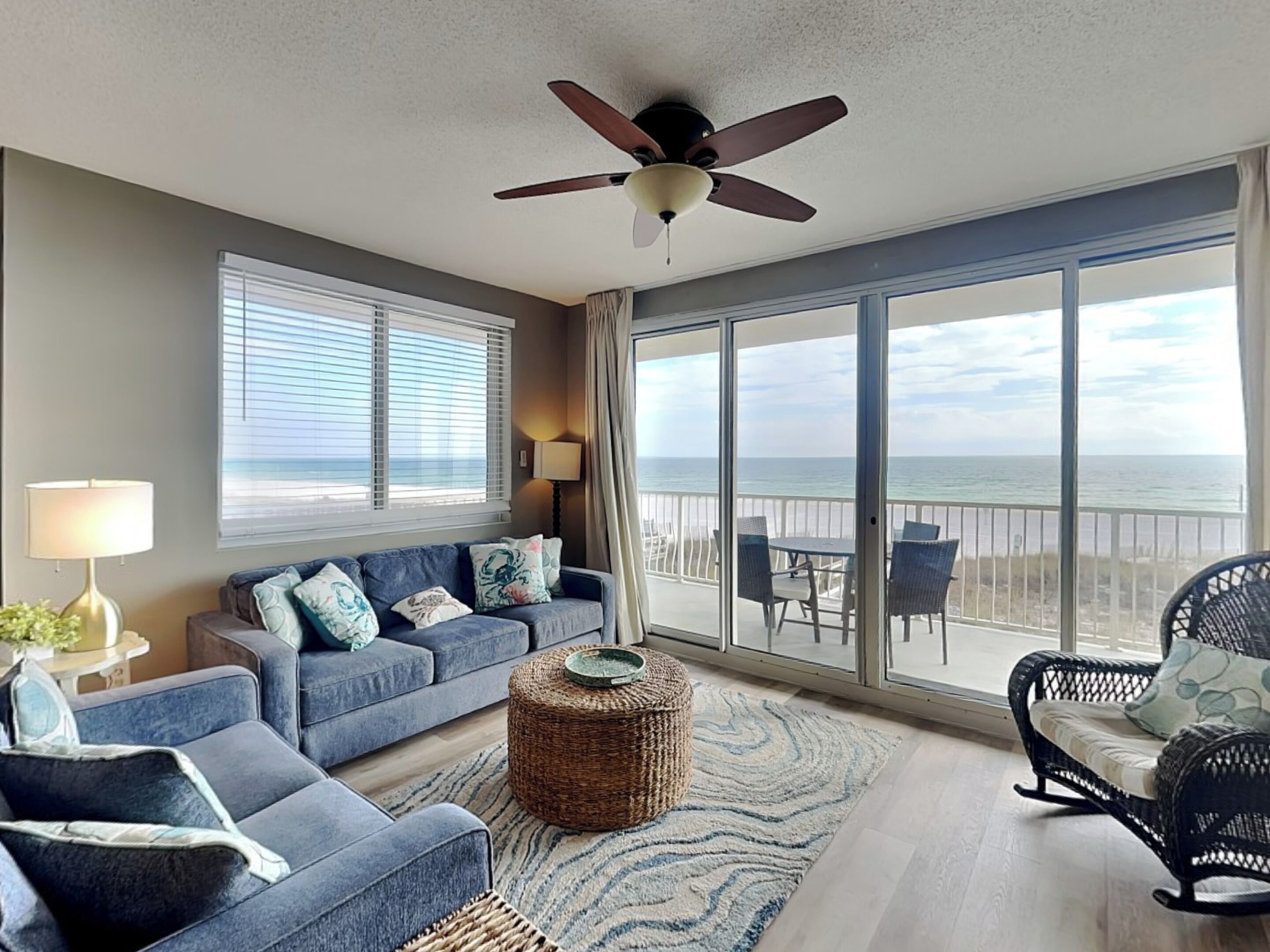 A spacious living room with a large window offering a view of the beach, a ceiling fan, and a cozy seating area with a wicker ottoman.