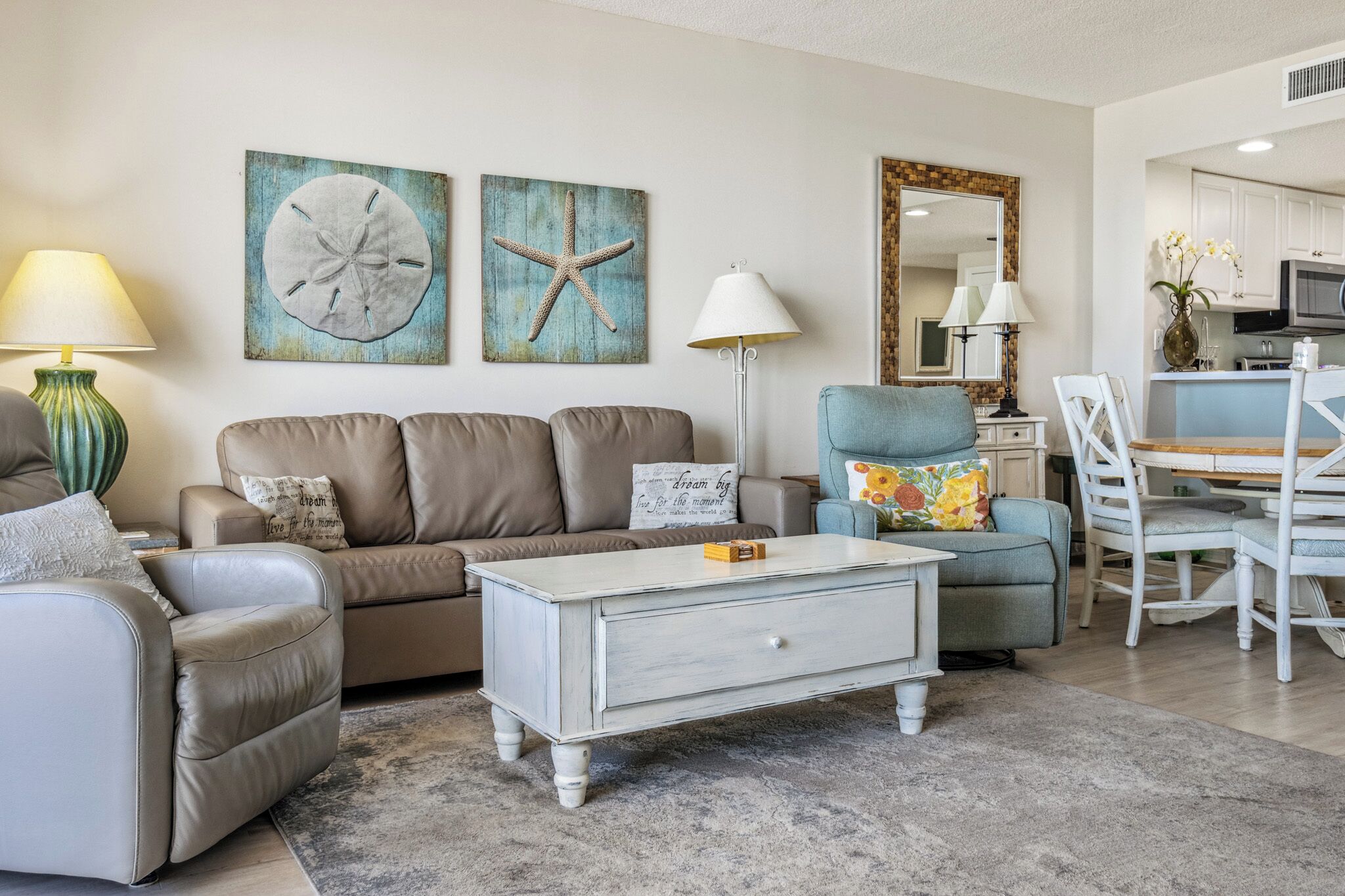 A living room with a beige sofa, a white coffee table, and a blue armchair. There are two wall art pieces, a lamp, and a mirror.