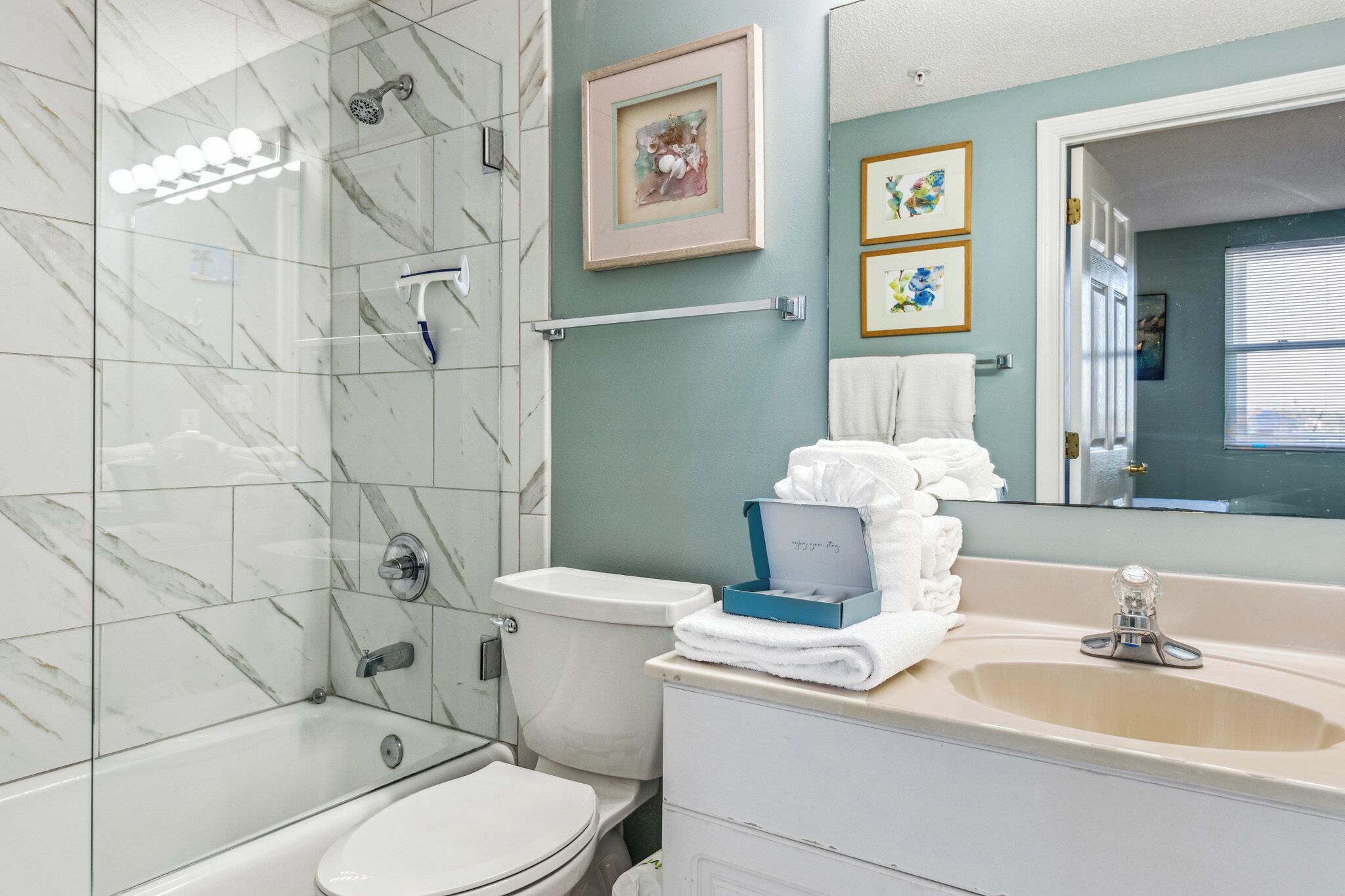 A modern bathroom with a glass-enclosed shower, a toilet, and a sink with a mirror above it. There are framed pictures on the wall and a towel rack with towels.