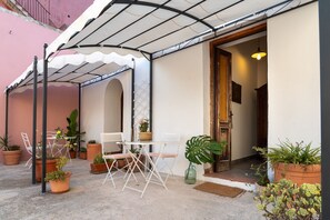 Een terras buiten met een tafel en stoelen, omringd door potplanten en een overdekte zithoek.
