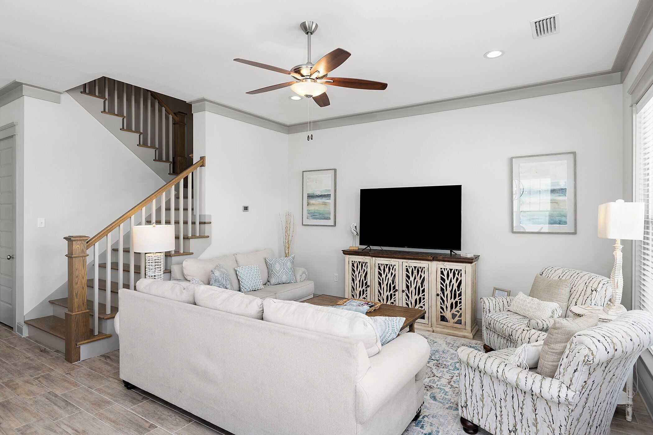 A living room with a staircase, a ceiling fan, a flat-screen TV, and a patterned armchair.