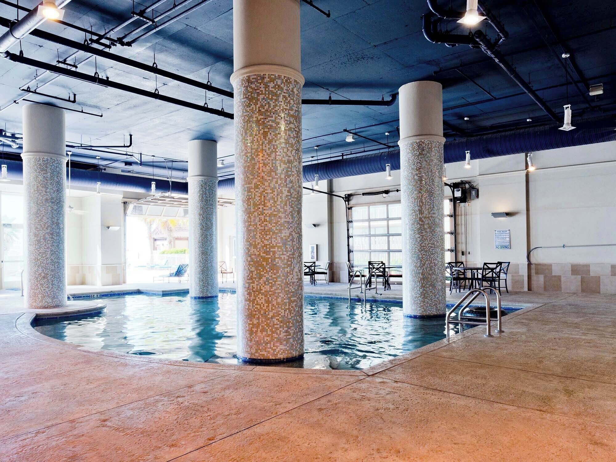 An indoor swimming pool with tiled columns and a tiled floor, surrounded by chairs and tables.