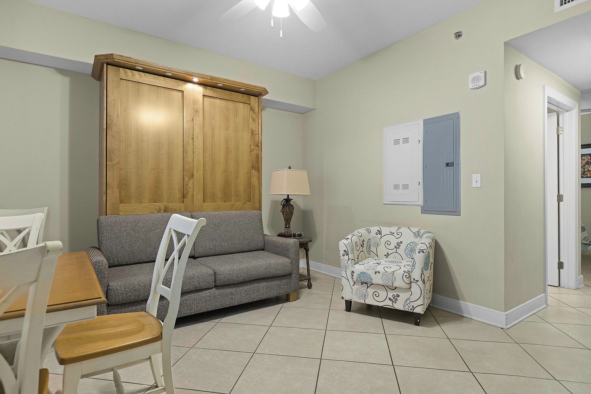 Une pièce comprenant une armoire en bois, un canapé gris, un fauteuil à motifs, une petite table avec une lampe et une chaise blanche.