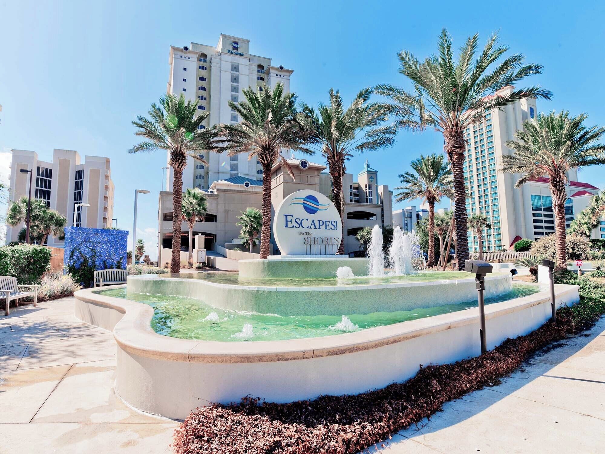 L’entrée d’un hôtel, avec une enseigne indiquant « Escapades ! En direction des rivages », et une fontaine au premier plan.