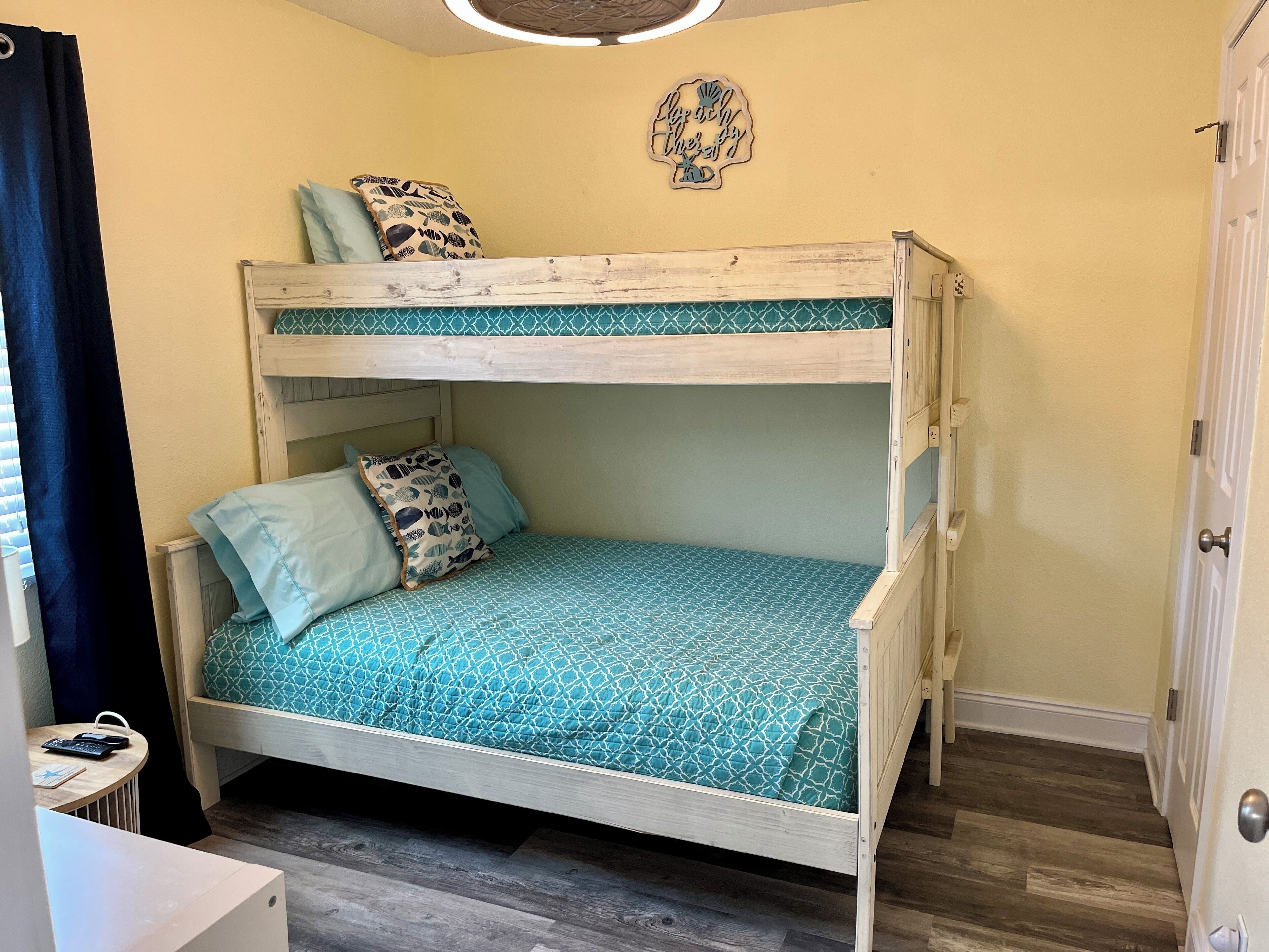 Una cama litera con una colcha estampada, una mesita con un bolso y un teléfono, y una decoración en la pared.