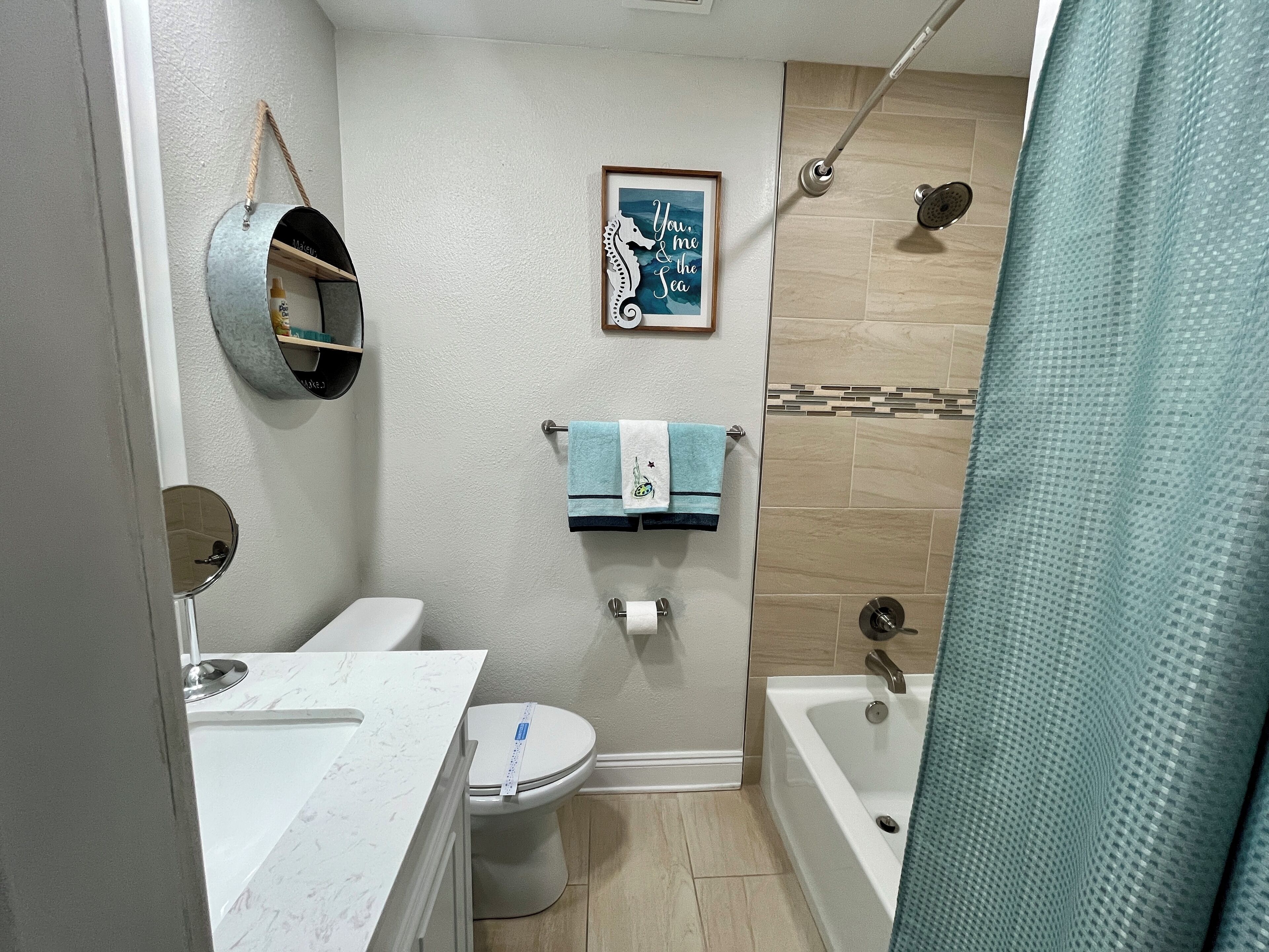 Un baño con inodoro, lavamanos y bañera. Hay una cortina de ducha, un perchero con toallas y un cuadro en la pared.