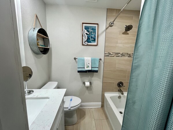 A bathroom with a toilet, sink, and bathtub. There is a shower curtain, a towel rack with towels, and a framed picture on the wall.