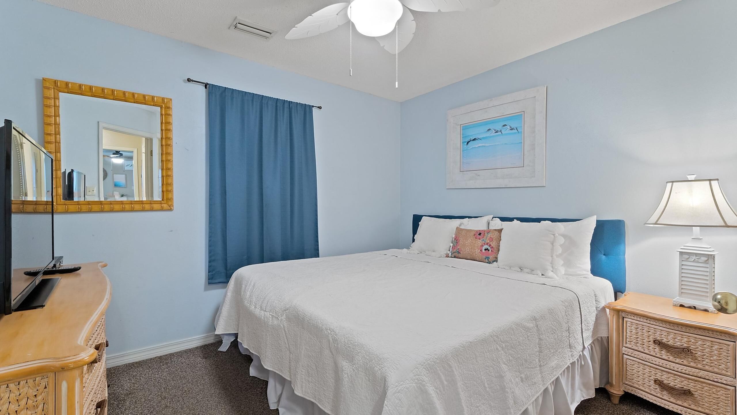 Une chambre à coucher avec un lit, une commode, un miroir, un ventilateur de plafond et un tableau accroché au mur.