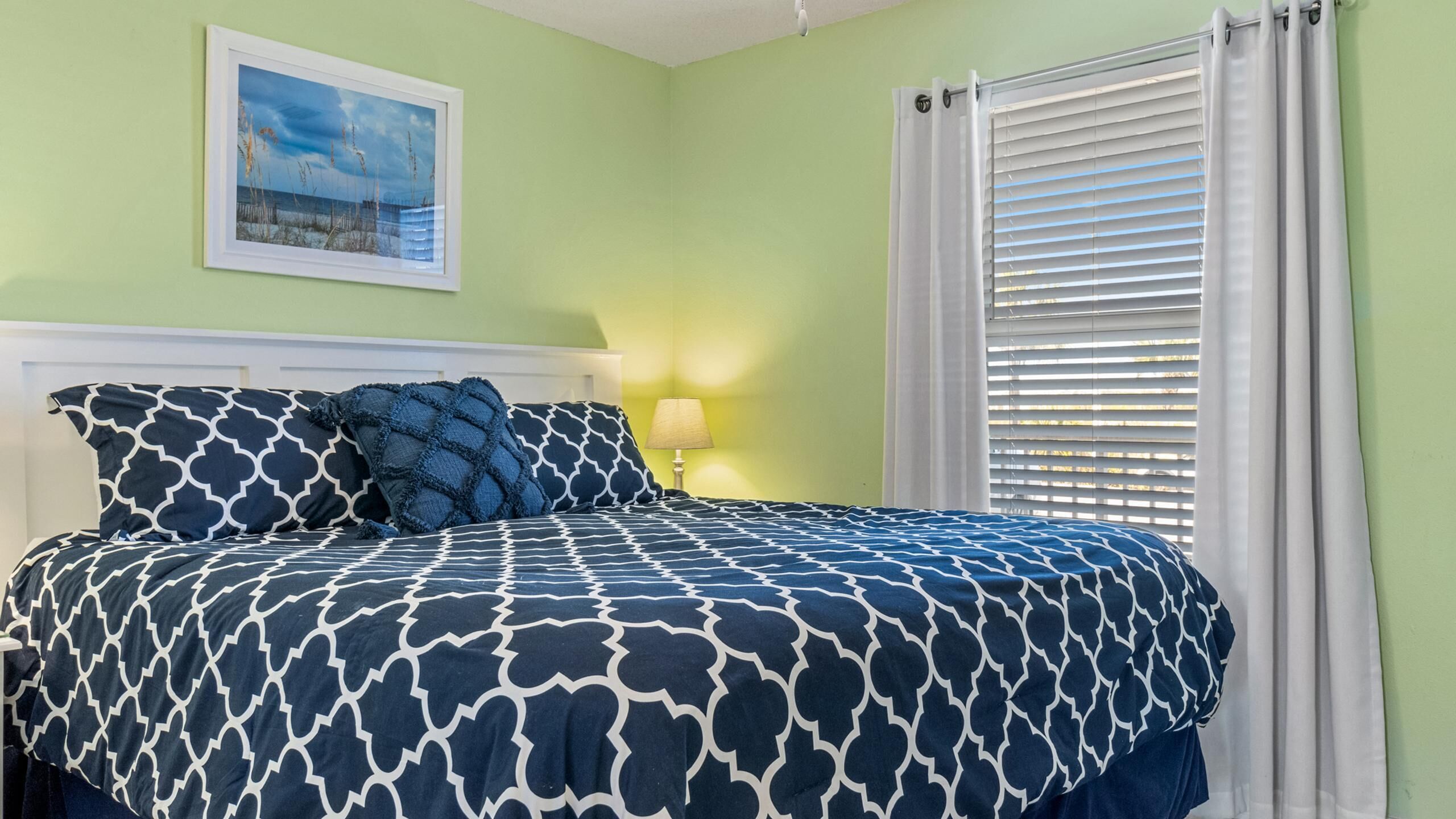 A bedroom with a bed, a nightstand, a lamp, a window with blinds, and a framed picture on the wall.