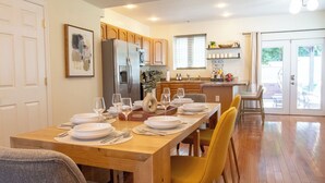 Un coin repas avec une table en bois dressée pour quatre personnes, une cuisine en arrière-plan et une vue sur l’extérieur par une porte vitrée.