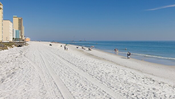 Ampia spiaggia sabbiosa con cielo azzurro limpido, edifici a sinistra e persone che passeggiano lungo la riva.