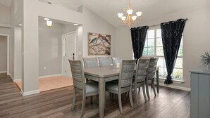 A dining area with a rectangular table, six chairs, a chandelier, and a window with curtains. - Happy Ours by Southern Vacation Rentals (Foley)