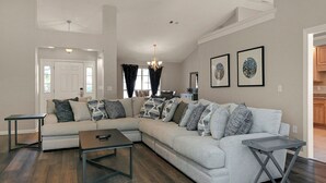 A living room with a sectional sofa, a coffee table, and a side table. There is a door leading to another room, a chandelier, and framed artwork on the wall. - Happy Ours by Southern Vacation Rentals (Foley)