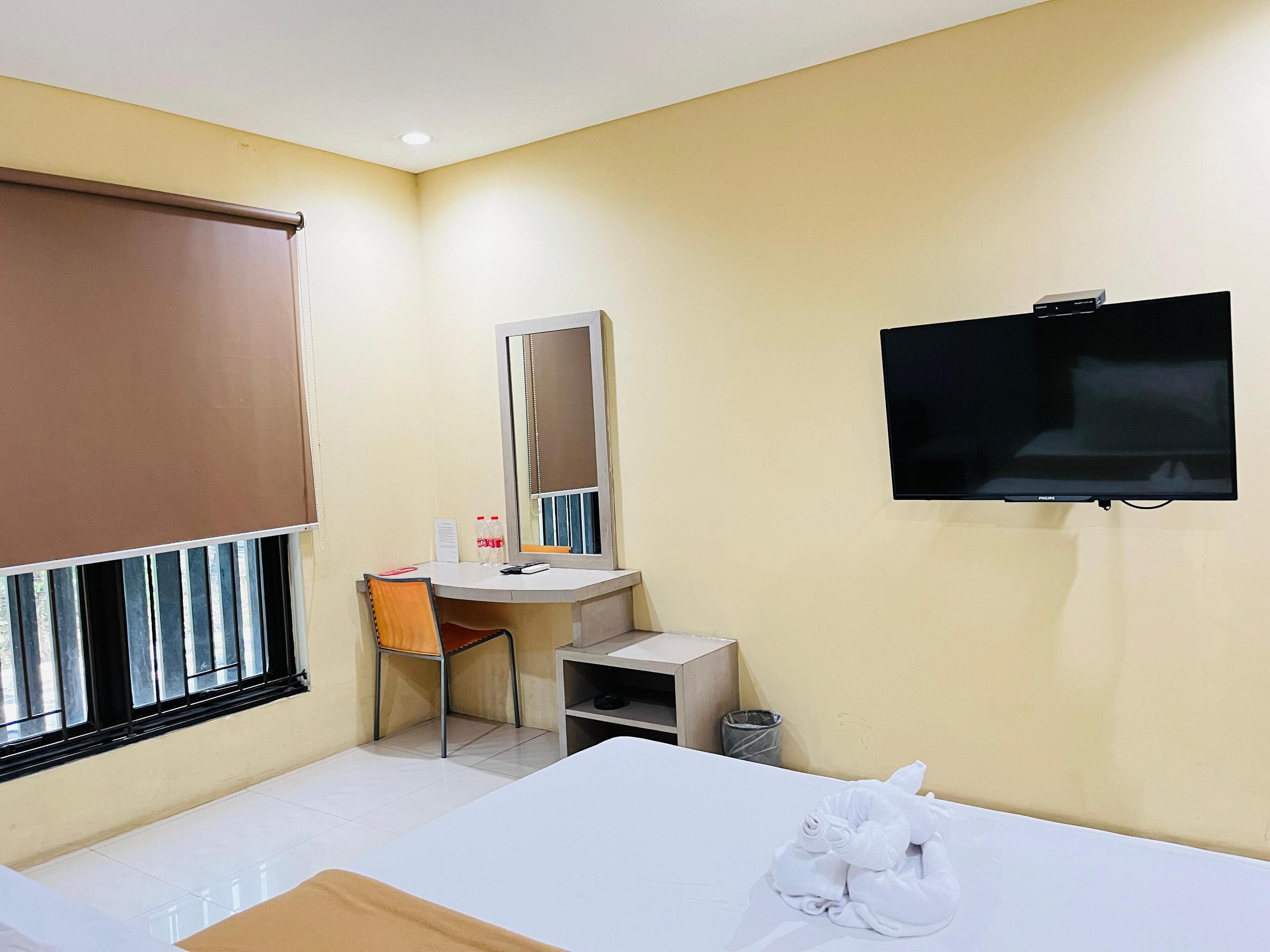 Habitación de hotel con cama, escritorio, silla, televisor, espejo y ventana con persianas.