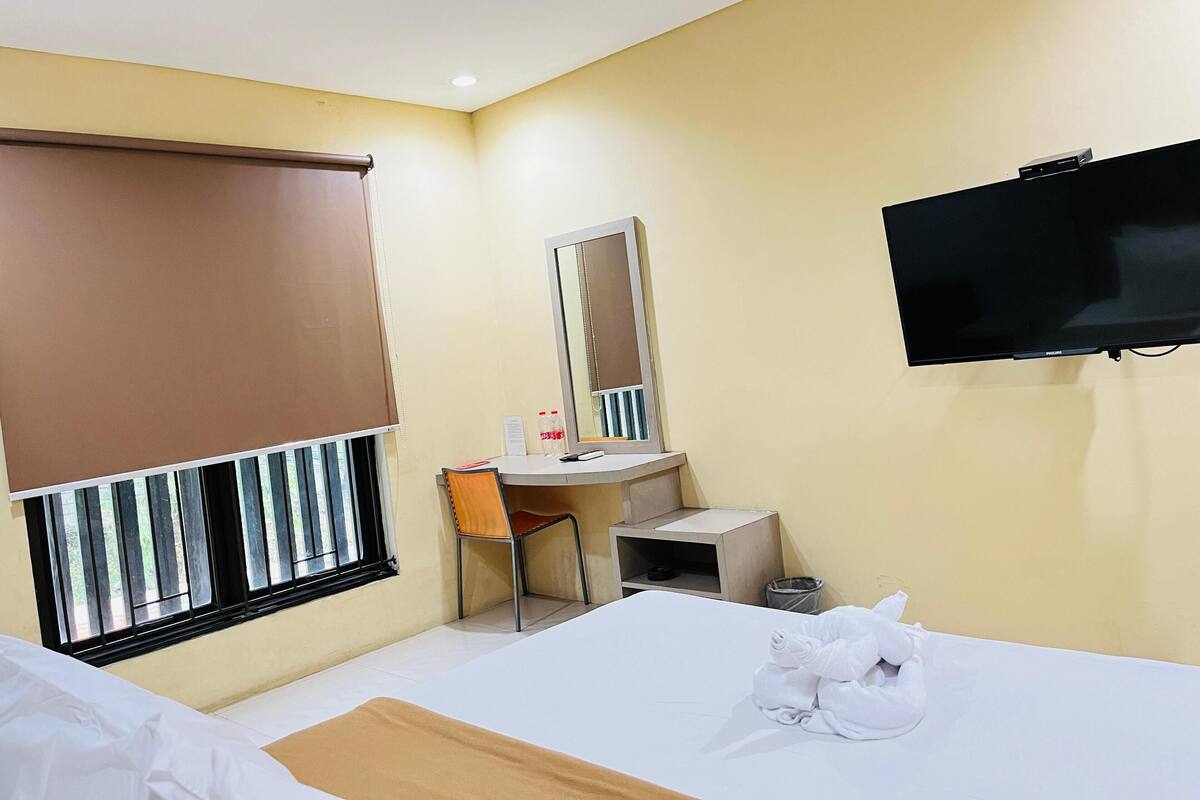 A hotel room with a bed, a desk with a chair, a mirror, a television, and a window with blinds.