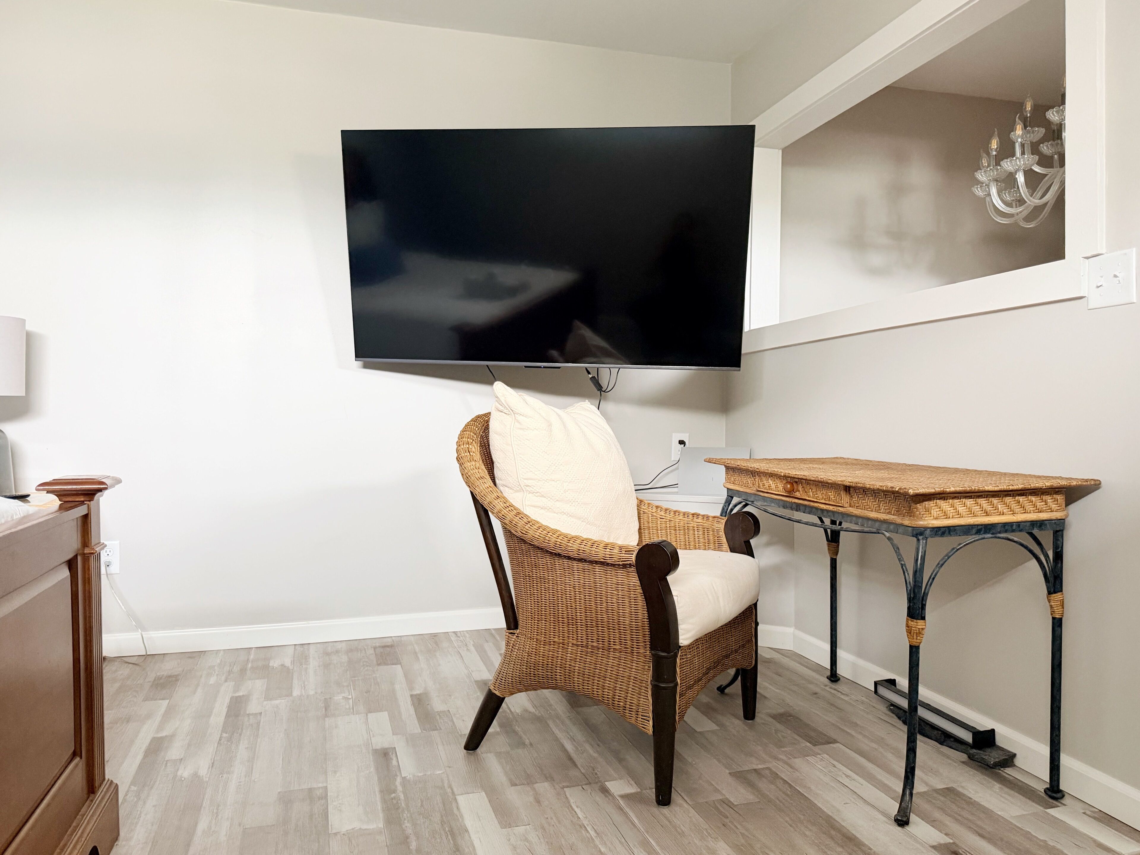 Une pièce dotée d’un téléviseur à écran plat fixé au mur, d’un fauteuil en osier avec un coussin, d’une petite table en bois et d’une commode en bois.
