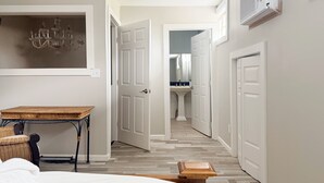 Un pasillo con una cama, una mesa y una silla, que conduce a un baño con lavamanos y espejo.