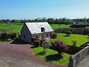 A stone farmhouse with a sloped roof, surrounded by a green field and a few trees.