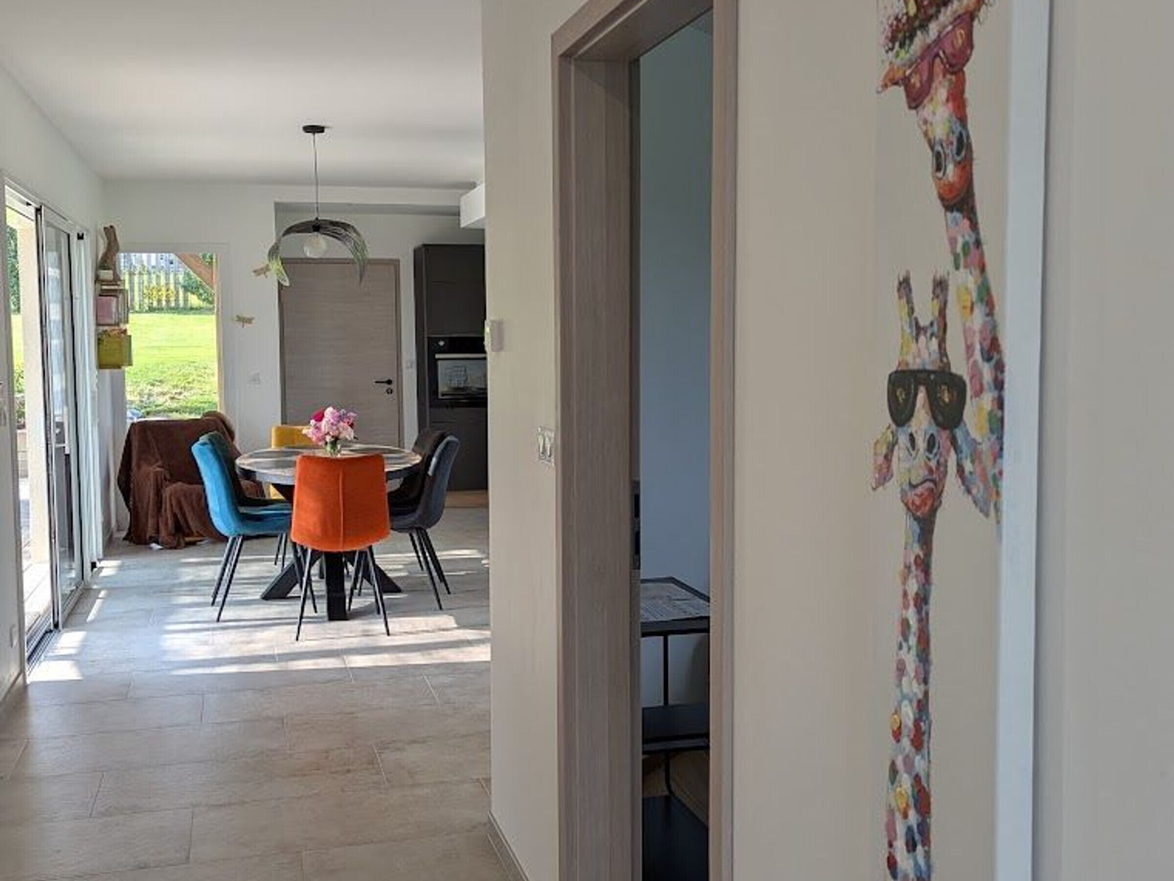 A modern dining area with a round table and chairs, a kitchen in the background, and a giraffe mural on the wall.